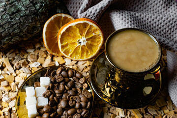 Aromatic dark coffee, with milk with bubbles on the surface in a dark gold Cup on a saucer, next to whole grains, peeled and dried oranges. Background of alder chips and wood snags in the background