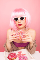 Stylish girl in a pink wig eats delicious donuts. Shot in a studio on a pink background.