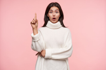 Excited young attractive brunette woman dressed in white woollen sweater keeping her forefinger...