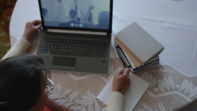 top view, aged female undergoes further learning, during an online schooling, raises her hand and writes answer in scratchpad while sitting in apartment