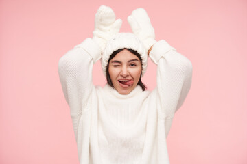 Funny shot of young lovely brown haired woman with casual hairstyle keeping raised hands under her head while winking cheerfully at camera and shwoing her tongue