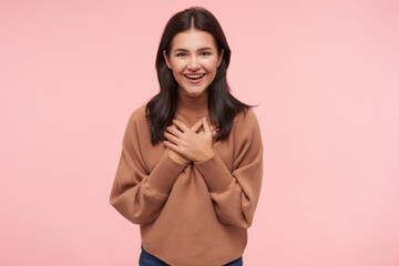 Happy young attractive brown haired woman keeping palms on her chest while looking cheerfully at camera with wide smile, standing over pink background