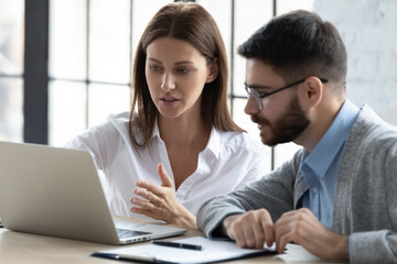 Confident businesswoman pointing at laptop screen, training new employee, helping with corporate software, serious mentor coach teaching worker, colleagues discussing online project, working together