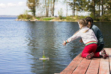 Fototapeta na wymiar Two little sisters are sitting on wooden walkways and letting a paper boat into the lake.