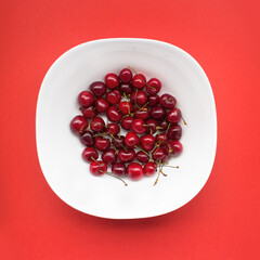 Cherry in a white plate on a red background. The composition of 
cherries on a colored background. Minimalism food