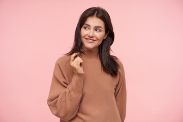 Fototapeta premium Cheerful young charming brunette female with natural makeup touching her hair with raised hand and looking gladly aside, standing over pink background