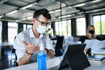 Young people with face masks back at work in office after lockdown, disinfecting hands.