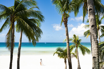 Fototapeta na wymiar (Selective focus) Stunning view of woman walking on a white sand beach bathed by a turquoise sea, beautiful coconut palm trees in the foreground. White Beach, Boracay Island, Philippines.