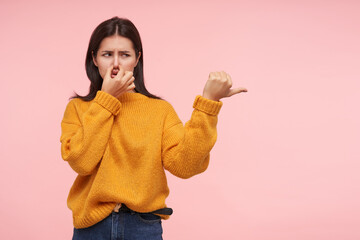 Displeased young brown haired woman dressed in yellow sweater closing her nose with fingers while...