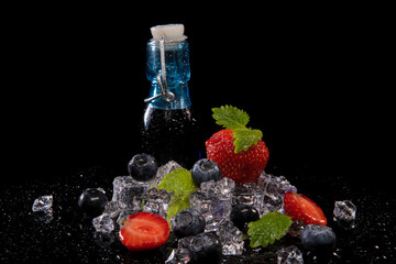 Strawberries, blueberries, mint leaves, ice cubes and a bottle of blackberry juice with drops of water on a black background with reflection