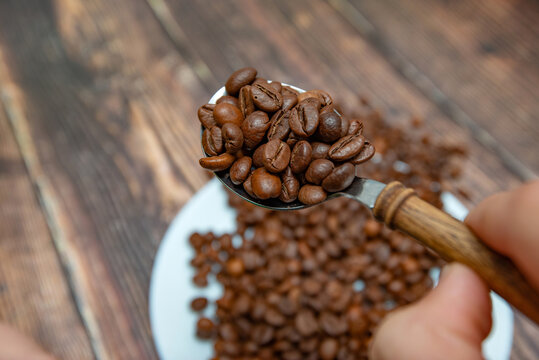 A man holds roasted coffee grains in a tablespoon on a background of a white plate and dark wood.