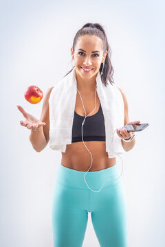 Slim beautiful girl in sports clothes smiles at the camera, with a towel around her neck a mobile phone in her hand and tossing in apple with the other hand on an isolated background.