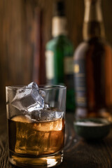 A glass of whiskey with ice, in the background are bottles on a wooden table of a bar counter, shallow depth of field, selective focus. The concept of alcoholic drinks in a roadside bar.