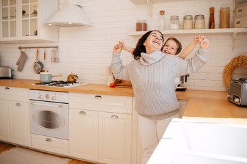 Mom with her son on the background of the kitchen are playing and hugging. A young woman and a little boy are laughing and smiling at home. Caring for the family, attention to the child.