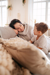 Young mother smiles at a child boy sitting on a bright sofa at home. Parent with son study drawing with felt-tip pens.