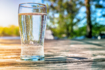 Naklejka na ściany i meble tall transparent glass with water on a wooden surface on a background of a summer landscape, blurred background