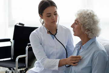 Young female physician caring about elderly disabled woman sit on couch using stethoscope listen...