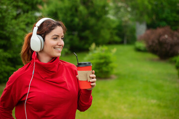 Red-haired woman listens to music on headphones and drinks coffee with a straw in the park. A girl enjoys a walk on a sunny summer day and holds a takeaway cup. earphones connected to a smartphone.