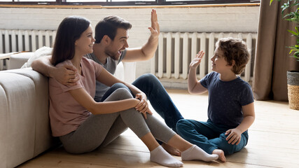 Happy young parents sit on warm wooden floor play have fun with excited preschooler son, smiling...