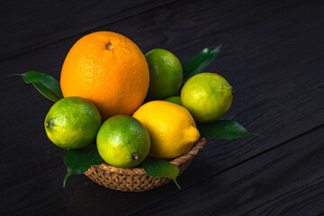 Orange, lemon and three limes lie in a tree plate on a dark brown table. Lots of citrus fruit, with lots of wits and useful amino acids