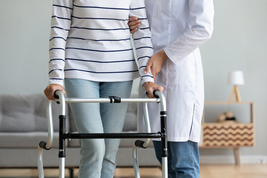 Cropped image caregiver helping to disabled aged female patient walk with walker physical therapist teach her, supporting during exercise therapy. Nursing at home, aftercare and physiotherapy concept
