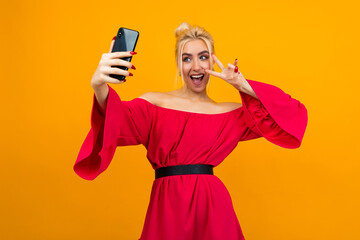 girl in an elegant red dress makes a photo of herself on the phone on a yellow studio background