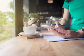 Fototapeta premium Young woman using smartphone while taking notes with cup of coffee on the wooden table in cafe.