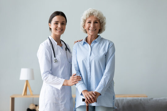 Elderly woman patient holds walking stick photo shooting with young caregiver practitioner in white coat portrait in living room, successful physiotherapy after accident, homecare and nursing concept