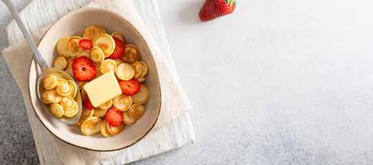 Pancake cereal banner. Mini pancakes in a bowl with strawberries and butter on white background. Trendy food during quarantine and lockdown. Top view, copy space. Menu restaurant. Kid's breakfast.