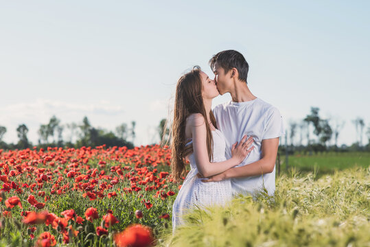 A couple of lovers in a poppy field, an Asian guy and a European girl hug and kiss on the border of wheat and poppy fields in the rays of the setting sun