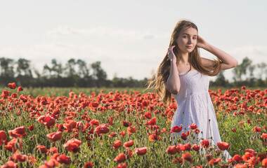 Fototapeta na wymiar A girl in a white sundress enjoys the warmth of the sun and walks through a poppy field