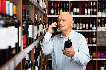 Senior man talking on mobile phone and choosing bottle of wine at the liquor store