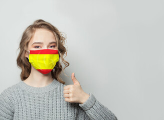 Happy woman in face mask holding thumb up with national flag Spain background. Flu epidemic and...