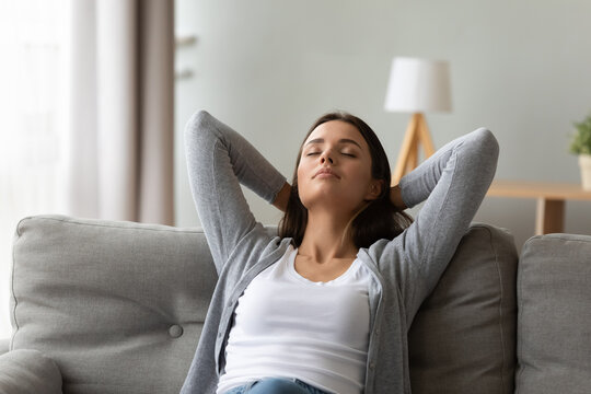 Calm 20s woman resting seated on couch in light warm living room put hands behind head enjoy fresh air-conditioned climate control, meditation, inner balance, relaxation at home free lazy day concept