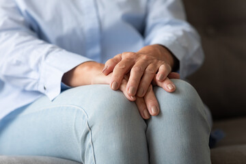 Close up image focus on folded female hands on knees, old woman sitting on couch has melancholic mood, lost in sad nostalgic thoughts, widow feels lonely, senile diseases, life health problems concept