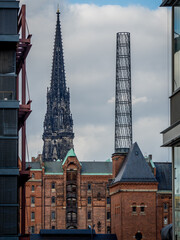 view of st nikolai memorial between modern office buildings and behind old speicherstadt warehouses next to the chimney of hafencity infocenter kesselhaus in front of cloudy sky