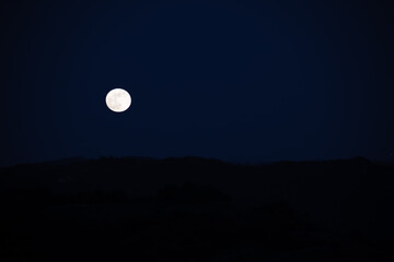 Huge super moon rising from behind the hills in a sweet spring night of may. Bright moon hovering the hills in the italian countryside. Visual poetry.