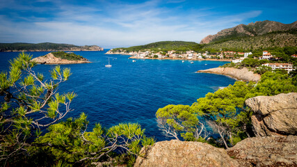 picturesque sight through pine tree branches over the bay of sant elm with boats in the middle, the island sa dragonera on the left side and a mountain ridge on the right side in the background