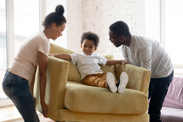 African couple carrying armchair where sit adorable small toddler son. Happy family placing...