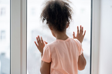Rear back view African little girl orphan standing near window alone looking outside feeling...