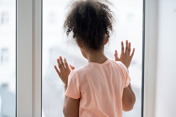 Rear back view African little girl orphan standing near window alone looking outside feeling loneliness. Adoption custody, social problems, dysfunctional family, parents divorce and orphanage concept