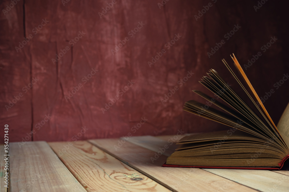 Poster beautiful ancient open old book on a red wooden table and dark-red wall background behind.