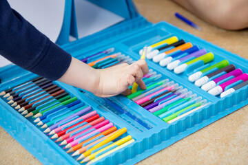 The child's hand takes a yellow felt-tip pen from a set of multicolored ones in a large blue box....