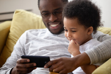 Close up American caring dad spend time with little son family sitting on sofa using smartphone watching cartoons having fun enjoy on-line app for kids. Parental control lazy weekends activity concept