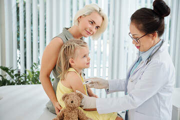 Pediatrician checking temperature of little girl sitting on bed in hospital ward with her mother