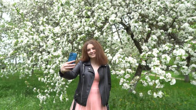 A girl in a dress is photographed in the garden. Photo session in a blooming garden. A beautiful girl is photographed on the phone.A girl in a dress takes a selfie. Lifestyle. 4K.