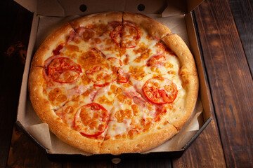 Heat the pizza in the box, pizza delivery. Pizza with ham, cheese, tomatoes, sauce on a wooden brown background. Empty space for text.
