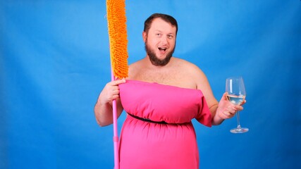 Funny bearded guy in a pink pillow dress with a mop and a glass of wine on a blue background. Crazy quarantine. Funny house cleaning. Fashion 2020. Put on a pillow. Challenge 2020 due to house