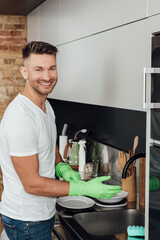 cheerful man wearing rubber gloves near white plates in kitchen
