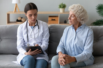 Young female caregiver medical worker in uniform providing care listen patient holds clipboard noting personal information writing health complaints of elderly woman seated on couch, nursing concept